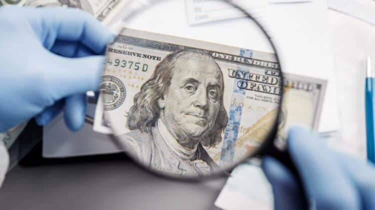 Gloved hands holding a magnifying glass over the face of Benjamin Franklin on a suspicious 100-dollar bill.