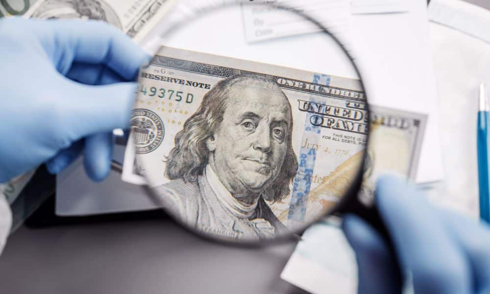 Gloved hands holding a magnifying glass over the face of Benjamin Franklin on a suspicious 100-dollar bill.